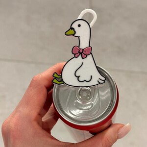 May include: A white goose-shaped soda can tab opener featuring a yellow beak, green feet, and a pink bow tie. The accessory is constructed with a white loop at the top and is designed to fit securely over the metal pull tab of a standard beverage can.