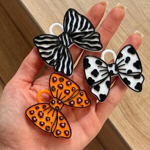 May include: Three bow-shaped charms with loops at the top. One charm features a black and white zebra stripe pattern, another has a white base with black cow spots, and the third is orange with a black and pink leopard print pattern.