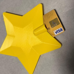 May include: A bright yellow star-shaped card holder with a central cylindrical slot. A gold-coloured Visa credit card is inserted into the slot. The star has a smooth, faceted surface and a distinct geometric design, providing a stable base for the card.