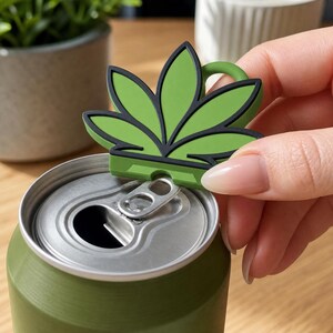 May include: A green and black leaf-shaped can opener is placed on top of a green can. The can opener has a loop for easy handling. The can opener is designed to open the top of a can.