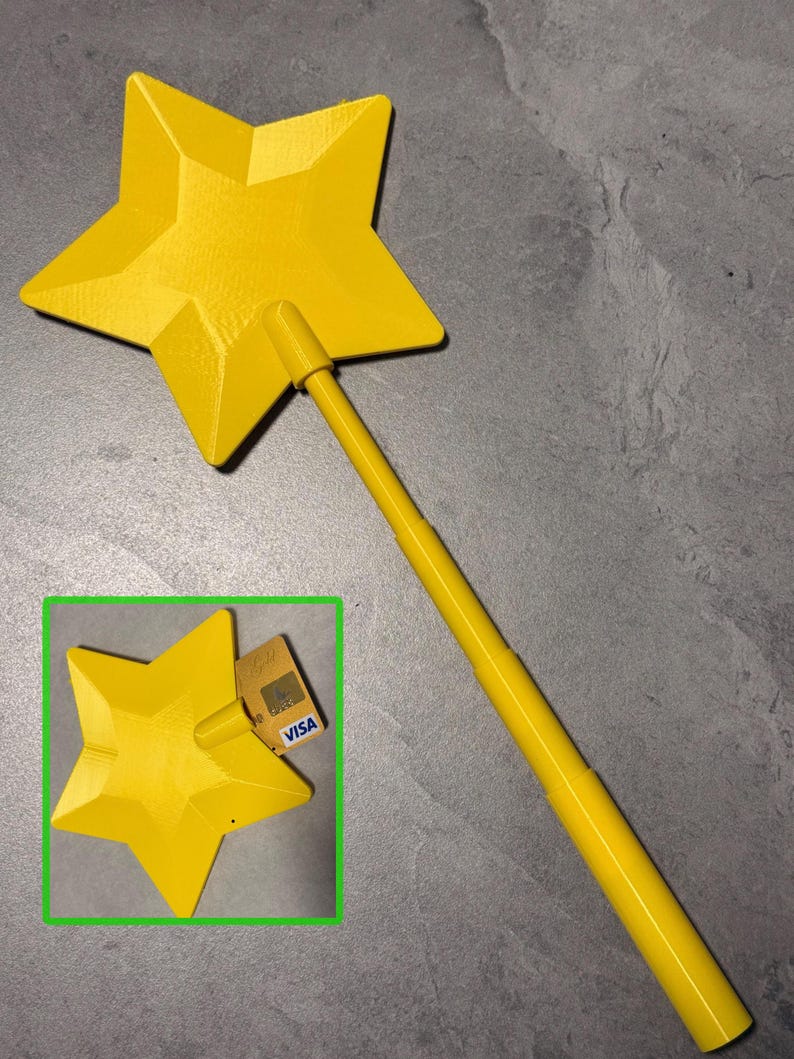 May include: A bright yellow star-shaped wand featuring a faceted, geometric design on the star head and a long, cylindrical handle. The inset image shows the same yellow star wand positioned next to a gold-coloured Visa credit card for scale.