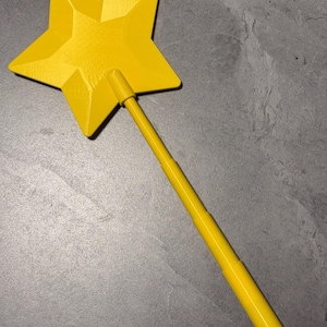 May include: A bright yellow star-shaped wand featuring a five-pointed star attached to a long, cylindrical handle. The star has a faceted, geometric surface texture, and the handle consists of multiple segments that appear to be connected in a linear fashion.