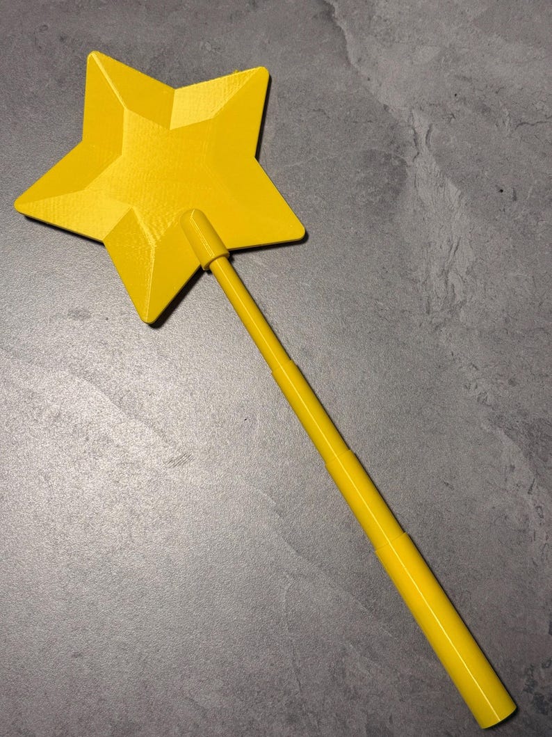 May include: A bright yellow star-shaped wand featuring a five-pointed star attached to a long, cylindrical handle. The star has a faceted, geometric surface texture, and the handle consists of multiple segments that appear to be connected in a linear fashion.