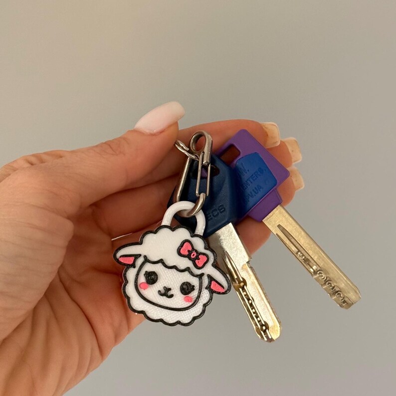 May include: A keychain with a white sheep design, featuring a pink bow and accents, hangs with several keys. The keys have blue and purple plastic heads and silver metal shafts. The keychain is held in a hand.