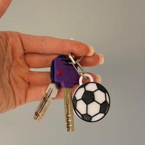 May include: A metal keyring featuring a round, black and white soccer ball charm attached to a silver split ring. The keyring also holds two keys, one of which has a purple plastic head with the text MULT-T-LOCK and a small red button.
