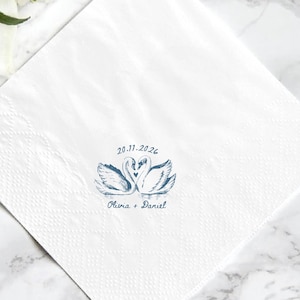May include: White square paper napkin with a blue swan design and the date 20.11.2026, along with the names Olivia + Daniel. The napkin is textured and appears to be made of paper.