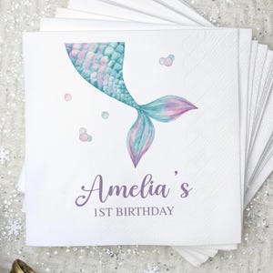 Puede incluir: Servilletas de papel blancas con un dise&ntilde;o de cola de sirena en tonos turquesa, rosa y morado. El texto "Amelia's 1st Birthday" est&aacute; impreso en una fuente cursiva. Las servilletas est&aacute;n apiladas en forma de abanico.