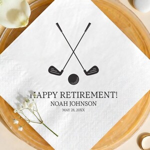 May include: White square napkin with a black golf club and ball graphic. The text "HAPPY RETIREMENT!" is printed below the graphic, along with a name and date. The napkin is on a gold-toned plate with decorative elements.