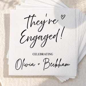 May include: A stack of square white paper napkins featuring black printed text that reads They're Engaged! with a small heart icon, followed by the word CELEBRATING and the names Olivia + Beckham in a decorative script font style.