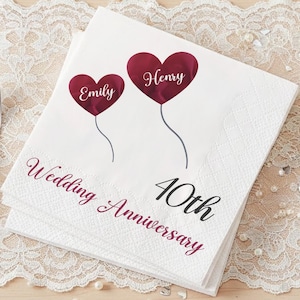 May include: White paper napkins with a burgundy heart design, featuring the names "Emily" and "Henry" and the text "Wedding Anniversary 40th". The napkins are stacked on a white lace tablecloth, with a fork and knife visible.