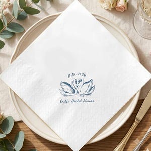 May include: A square white paper napkin featuring a blue ink illustration of two swans forming a heart shape with their necks. Above the swans is the date 11.15.2026, and below the swans is the text Emily's Bridal Shower in blue script.