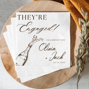 May include: White paper napkins with the text "THEY'RE Engaged!" and a champagne bottle illustration. The napkins also include the names "Olivia + Jack" and the date "20.10.2025".
