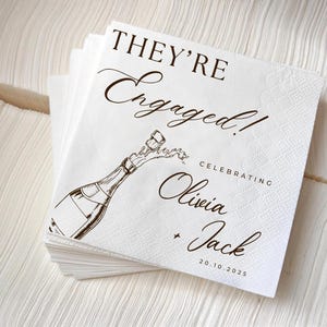 May include: A stack of white paper napkins with a brown illustration of a champagne bottle popping. The text reads "THEY'RE Engaged!" and "Celebrating Olivia + Jack 20.10.2025".