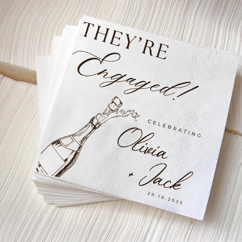 May include: A stack of white paper napkins with a brown illustration of a champagne bottle popping. The text reads "THEY'RE Engaged!" and "Celebrating Olivia + Jack 20.10.2025".