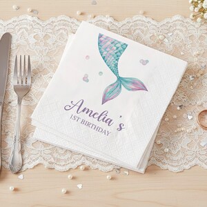 Puede incluir: Servilletas de papel blancas con un dise&ntilde;o de cola de sirena en tonos azules, rosas y morados. Las servilletas est&aacute;n personalizadas con el nombre "Amelia's" y "1ST BIRTHDAY" en una fuente cursiva. Las servilletas est&aacute;n sobre un mantel de encaje.