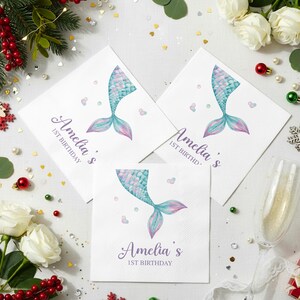 Puede incluir: Servilletas de papel blancas con un dise&ntilde;o de cola de sirena en tonos azules y morados. El texto "Amelia's 1st Birthday" est&aacute; impreso. Las servilletas est&aacute;n rodeadas de elementos decorativos, incluyendo flores y adornos.