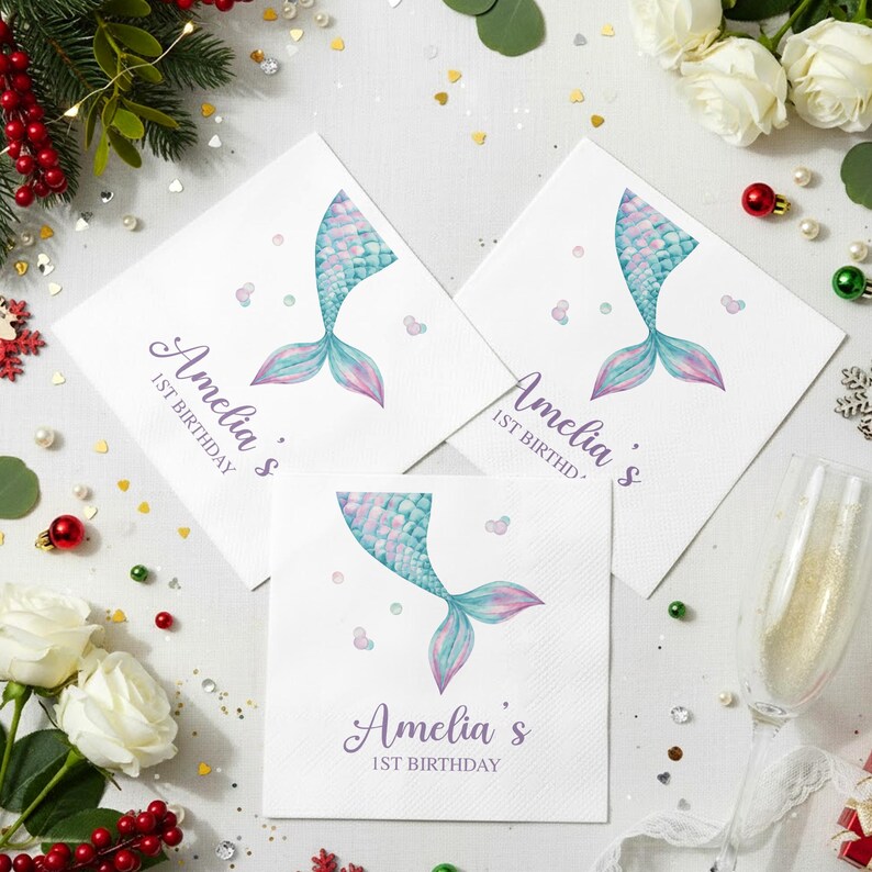 Puede incluir: Servilletas de papel blancas con un dise&ntilde;o de cola de sirena en tonos azules y morados. El texto "Amelia's 1st Birthday" est&aacute; impreso. Las servilletas est&aacute;n rodeadas de elementos decorativos, incluyendo flores y adornos.
