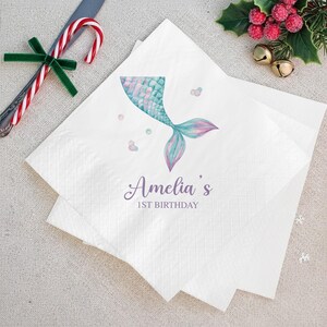 Puede incluir: Servilletas de papel blancas con un dise&ntilde;o de cola de sirena en acuarela en tonos azules, turquesas y rosas. El texto "Amelia's 1st Birthday" est&aacute; impreso en una fuente cursiva. Decoraciones festivas en el fondo.