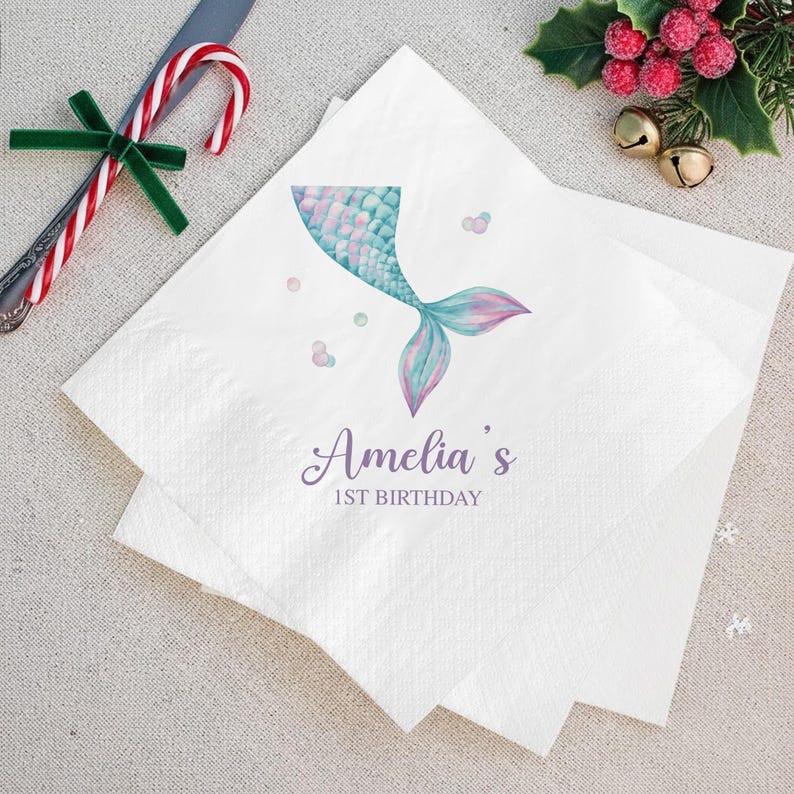 Puede incluir: Servilletas de papel blancas con un dise&ntilde;o de cola de sirena en acuarela en tonos azules, turquesas y rosas. El texto "Amelia's 1st Birthday" est&aacute; impreso en una fuente cursiva. Decoraciones festivas en el fondo.