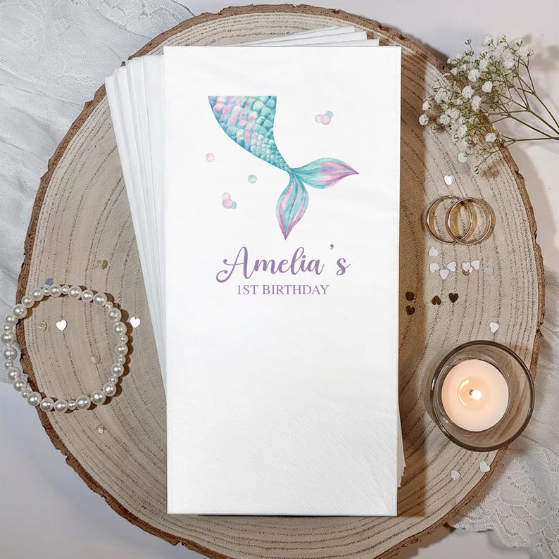 Puede incluir: Servilletas de papel blancas con un dise&ntilde;o de cola de sirena en acuarela en tonos turquesa y morado. El texto "Amelia's 1st Birthday" est&aacute; impreso debajo. Las servilletas est&aacute;n sobre una tabla de madera.