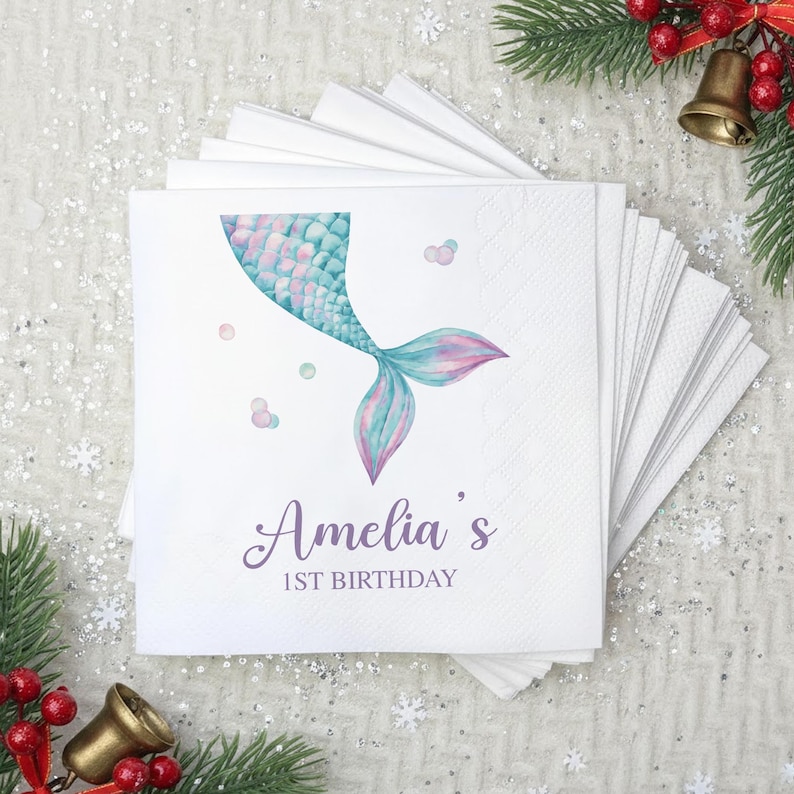 Puede incluir: Servilletas de papel blancas con un dise&ntilde;o de cola de sirena en tonos turquesa, rosa y morado. El texto "Amelia's 1st Birthday" est&aacute; impreso en una fuente cursiva. Las servilletas est&aacute;n apiladas en forma de abanico.