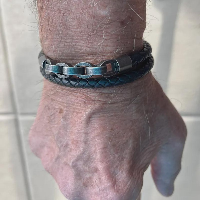 May include: A multi-strand bracelet featuring a dark braided leather cord paired with a metallic chain composed of interconnected oval links with a brushed silver finish and a smooth, dark grey cylindrical clasp connector.
