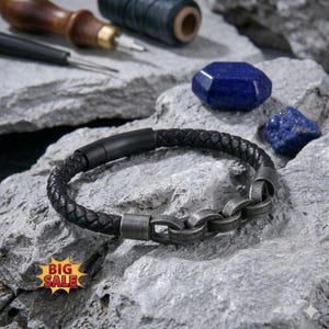 May include: A black braided leather bracelet featuring a matte black cylindrical clasp and a series of interconnected, weathered silver-toned metal links. The bracelet is displayed on a textured gray stone surface with blue stones.