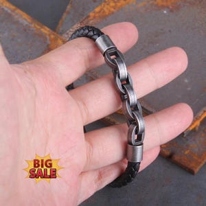 May include: A bracelet featuring a black braided leather cord and a series of interconnected, weathered silver-toned metal links. The design includes smooth cylindrical metal end caps that secure the leather cord to the central chain section.