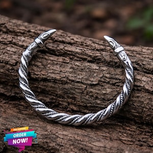 Könnte beinhalten: Ein silberfarbenes, verdrehtes Metallarmband mit spitzen Enden. Das Armband liegt auf einer strukturierten, braunen Holzoberfläche. Das Design weist ein geflochtenes, seilartiges Muster entlang des Bandes mit dekorativen Details an den Spitzen auf.