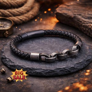 May include: A braided black leather bracelet featuring a matte black cylindrical clasp and a series of three interconnected, weathered silver-toned metal chain links. The bracelet is displayed on a dark, circular stone surface.