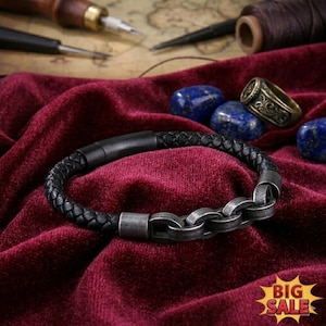May include: A black braided leather bracelet featuring a matte black cylindrical clasp and a series of interconnected, weathered silver-toned metal links. The bracelet is displayed on a deep red velvet fabric alongside several polished blue stones.