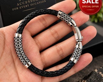 May include: A black braided leather bracelet featuring polished silver-tone metal accents with a textured, woven pattern. The bracelet includes a secure metal clasp mechanism and is presented against a dark box with gold text.