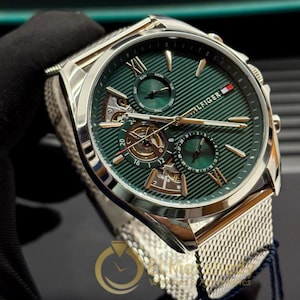 Puede incluir: Un reloj Tommy Hilfiger plateado con esfera verde y correa de malla. La esfera del reloj presenta agujas e indicadores dorados, con múltiples subesferas y el nombre de la marca. El reloj se muestra sobre un fondo oscuro.