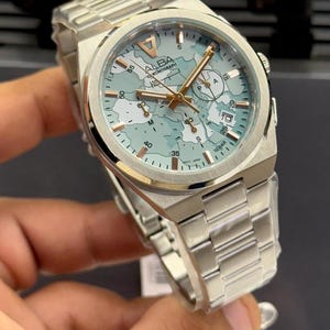 Op de afbeelding: Zilverkleurig Alba chronograaf horloge met een lichtblauwe wijzerplaat met een kaartachtig ontwerp en goudkleurige wijzers en markeringen. Het horloge heeft een roestvrijstalen band en een ronde wijzerplaat met de tekst "ALBA CHRONOGRAPH".