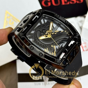 May include: Black Guess watch with a square face and a black strap. The watch face features a detailed design with gold-colored hands and the Guess logo. The watch has a unique, textured bezel.