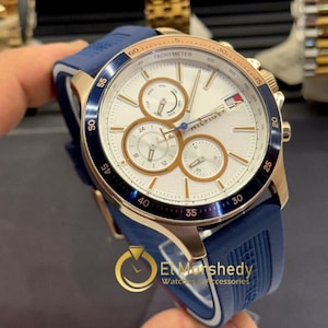 Könnte beinhalten: Eine Tommy Hilfiger Uhr mit weißem Zifferblatt, goldenen Akzenten und einem blauen Silikonarmband. Die Uhr verfügt über mehrere Hilfszifferblätter und eine Tachymeterlünette. Die Uhr wird von einer Person gehalten.