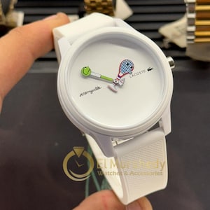 May include: White Lacoste watch with a tennis-themed design. The watch face features a tennis ball and racket graphic, with the word "Jeremyville" and the Lacoste logo. The watch has a white strap and a round face.