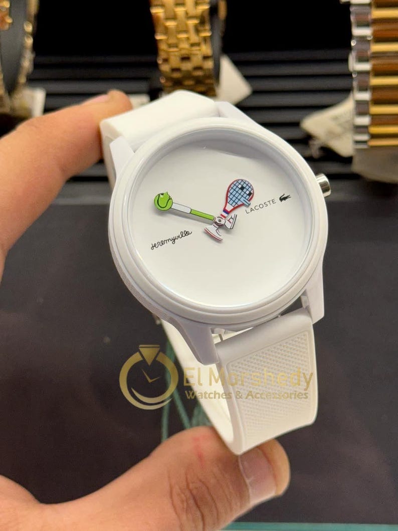 May include: White Lacoste watch with a tennis-themed design. The watch face features a tennis ball and racket graphic, with the word "Jeremyville" and the Lacoste logo. The watch has a white strap and a round face.