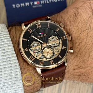 May include: A Tommy Hilfiger watch with a black face, gold-tone detailing, and a brown leather strap. The watch has Roman numeral hour markers and several sub-dials. The watch is shown on a wrist.