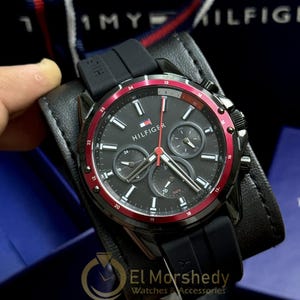 Op de afbeelding: Zwarte Tommy Hilfiger horloge met een rode lunette en zwarte rubberen band. De wijzerplaat van het horloge is zwart met zilveren wijzers en markeringen. Het horloge heeft drie sub-wijzerplaten en het Hilfiger-logo.