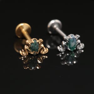 May include: Two flat-back labret studs featuring a frog-shaped design. One stud is gold-toned and the other is silver-toned. Each frog has a small, oval-shaped, dark green moss agate gemstone set into its back, with detailed legs and head.