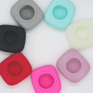 May include: A selection of colourful silicone bottle stoppers in shades of red, pink, black, grey, light blue, yellow, and lavender. Each stopper has a square shape with a circular opening in the centre.