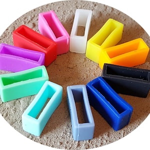 Puede incluir: Colección de pasadores de silicona para correas de reloj en varios colores: rojo, rosa, morado, verde, azul, gris, amarillo, naranja, negro y blanco. Los pasadores, de forma rectangular con una abertura central, están dispuestos sobre una superficie texturizada de color marrón claro.