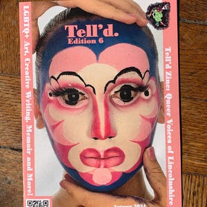 Op de afbeelding: Een magazinecover met een roze en blauw ontwerp met een gezicht met theatrale make-up. Het tijdschrift heet "Tell'd. Edition 6" en bevat tekst over LGBTQ+ kunst, creatief schrijven en memoires. De cover heeft ook een QR-code en de tekst "Herfst 2024."