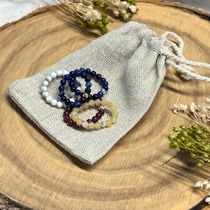 May include: A collection of beaded bracelets in white, navy blue, burgundy, and yellow, displayed on a linen pouch. The bracelets are made of small, round beads. The pouch is tied with a natural rope. The background is a wooden surface.
