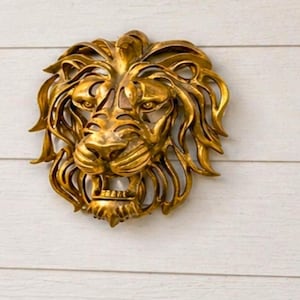 Op de afbeelding: Een gouden leeuwenkop-wandsculptuur met gedetailleerde manen en gezichtskenmerken. Gemonteerd op een witte, horizontaal geplankte muur, geschikt voor buiten- of entree-decoratie. De bek van de leeuw is lichtjes geopend.