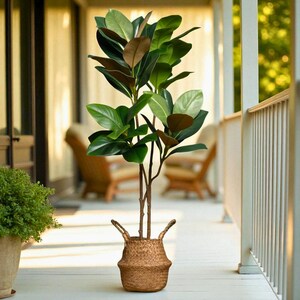 以下が含まれることがあります： 大きな緑と茶色の葉を持つ人工植物。持ち手付きの織りバスケットに入っています。白いベランダにあり、手すりと背景に椅子があります。植物の高さは約150cmです。