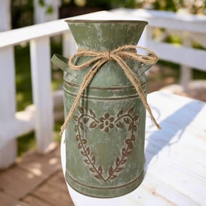 Może przedstawiać: Postarzany zielony metalowy wazon z motywem serca i kwiatów. Wazon jest zawiązany naturalną kokardą ze sznurka. Wazon ma uchwyt po każdej stronie i stoi na białej powierzchni.