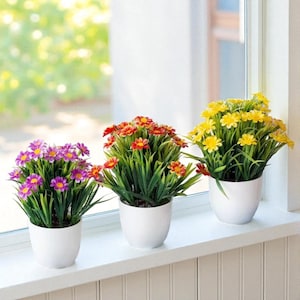 May include: Three potted artificial flowers in white ceramic pots. The flowers are purple, red-orange, and yellow, with green foliage. The pots are arranged on a white windowsill, with a blurred green background.