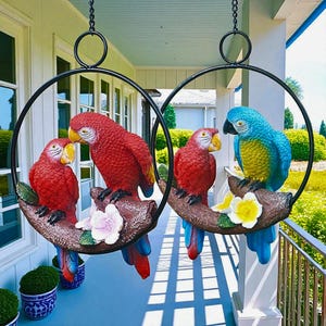 May include: Two hanging parrot sculptures. One features two red parrots, the other has a red parrot and a blue and yellow parrot. Each parrot is perched on a brown branch with flowers, suspended by a black metal ring.